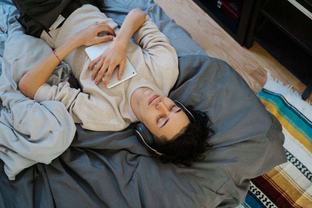 Image of a person lying on bed with headphones on, eyes closed, and a tablet held to their chest, depicting a mood of relaxation.