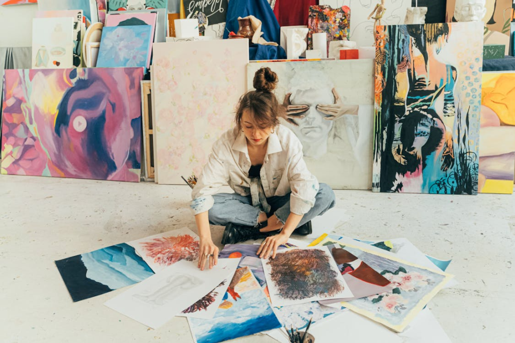 Woman sitting on the floor surrounded by scattered paintings with a wall of paintings in the background.