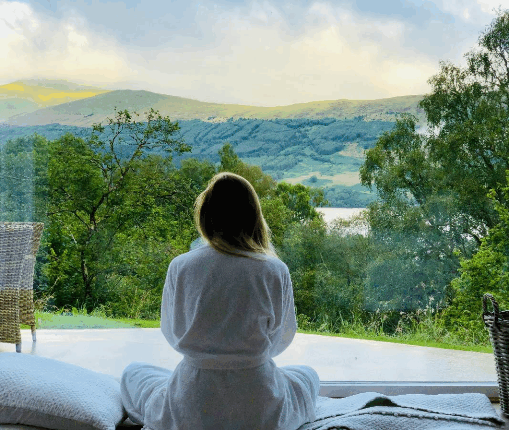 Woman in a robe sitting by a window, gazing at mountains and trees.