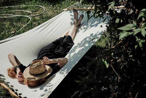 Person lying in a hammock outdoors with a hat covering their face, resting in the shade.