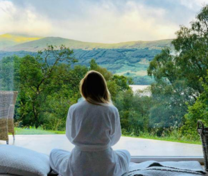 Woman in a robe sitting by a window, gazing at mountains and trees.