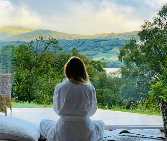 Woman in a robe sitting by a window, gazing at mountains and trees.