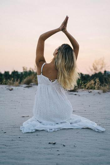 Woman meditating on a sandy beach in a white dress at sunset.
