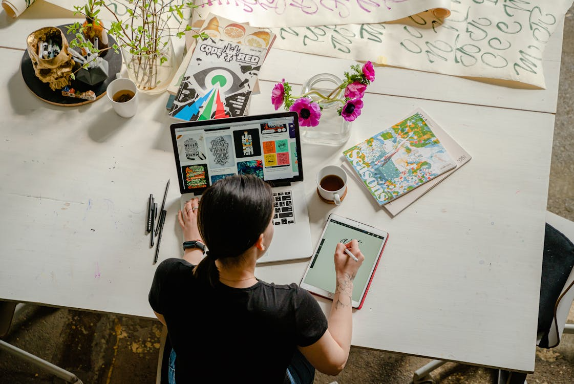 Aerial image of a person using a laptop and a tablet to sketch, with a coffee cup, books, and pens randomly lying around her on the table.