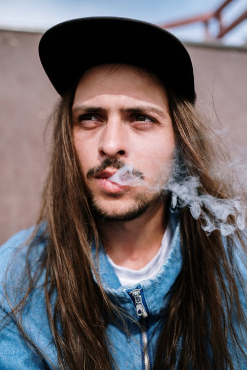 Close-up of a man with long hair releasing smoke from his mouth.