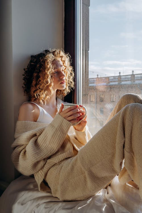 A woman in a sweater sits by a window holding coffee in soft natural light