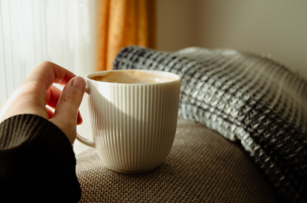 A hand holding a white mug in a cozy indoor setting with sunlight streaming through a window