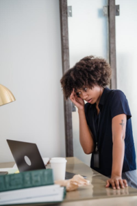 A tired businesswoman at a desk with a laptop, holding glasses and showing signs of stress