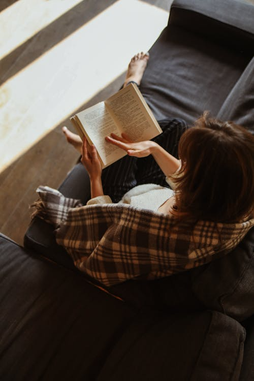  A woman sitting on a sofa reading a book with a plaid blanket in soft sunlight