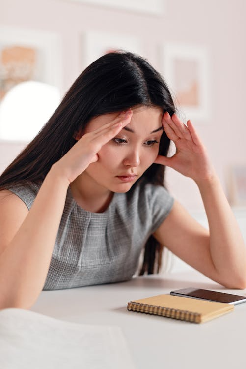 A woman sitting at a desk with both hands pressed on top of her head, appearing overwhelmed and mentally drained.