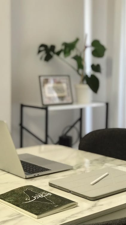 A clean, modern workspace featuring a laptop, a spiral notebook, and a small plant arranged on a white marble surface.