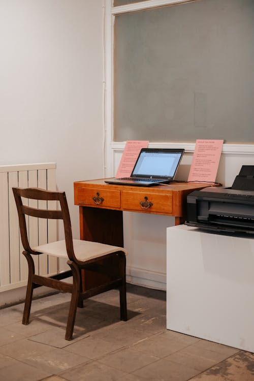 A minimalist workspace featuring a wooden chair, a wooden desk, an open laptop, and a compact printer in a simple indoor room.
