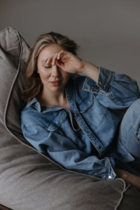 A woman in a casual jacket sitting on a chair with her eyes closed and head resting on her hand in front of a gray background.