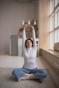 A woman seated on a mat in a calm indoor setting with her arms raised upward in a gentle yoga stretch.