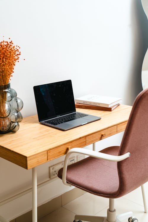A modern minimalist desk setup with a laptop, stacked books, a small decorative vase, and a lamp in a tidy indoor workspace.