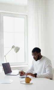 A man in glasses working at a bright, organized home office desk with a laptop and a coffee cup nearby.