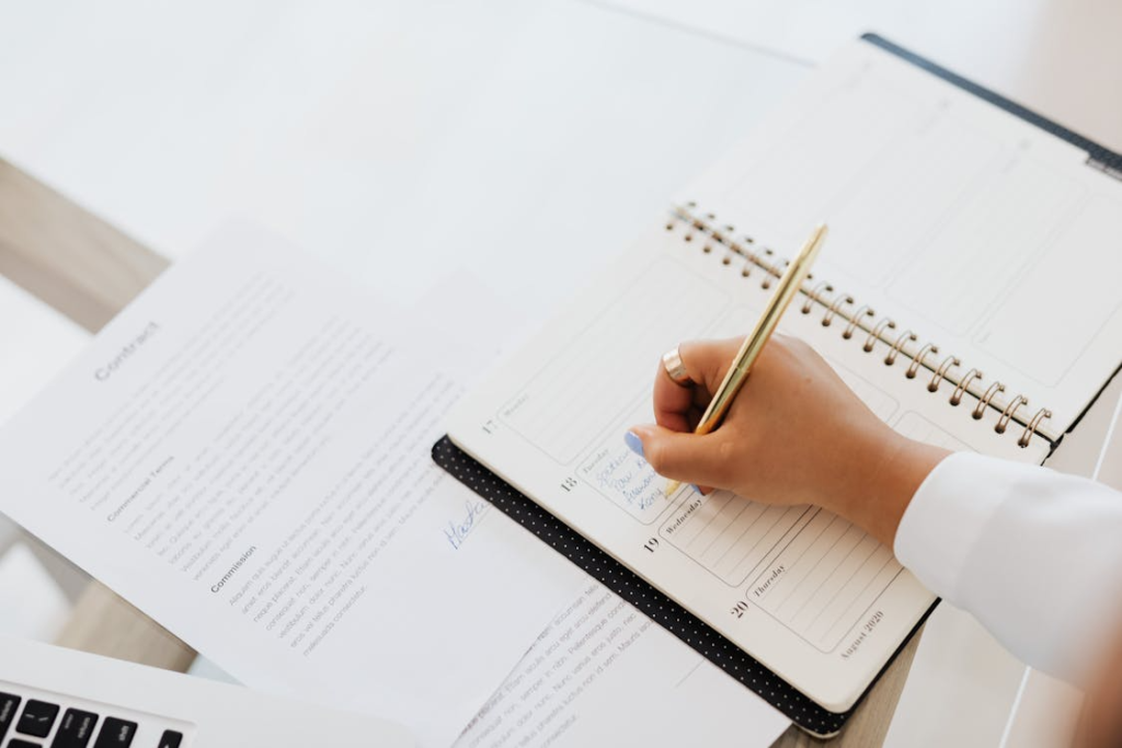 A hand writing in an open spiral notebook with a gold pen on a clean white desk surface.