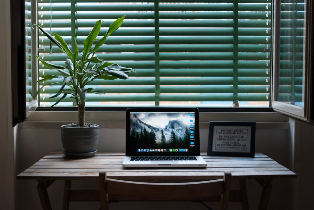 A MacBook Air resting on a wooden desk beside a small potted plant and a framed picture near a window.