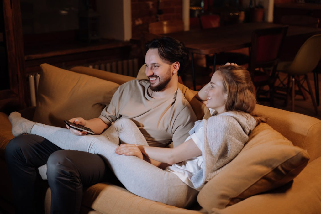 A man and woman sitting close together on a sofa in a warmly lit living room during the evening.