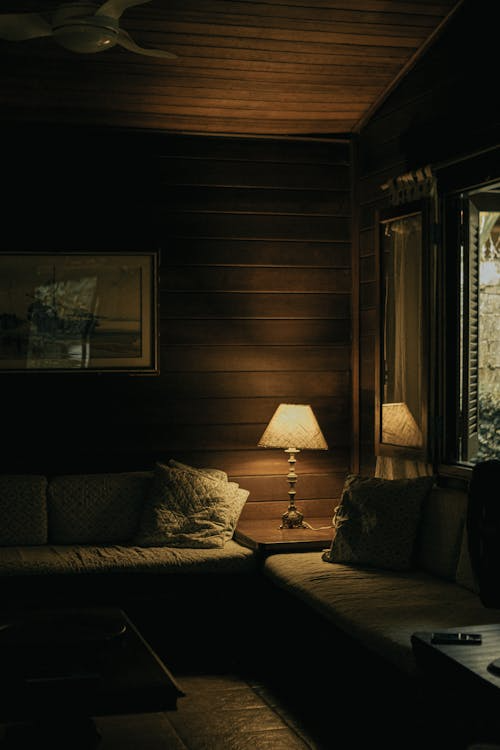 A rustic cabin living room with a warm-lit lamp, wood-paneled walls, soft cushions, and a peaceful evening atmosphere.