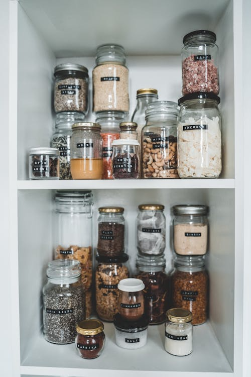 A neat row of clear labeled glass jars on a white wooden shelf, each containing different dry goods and pantry staples.