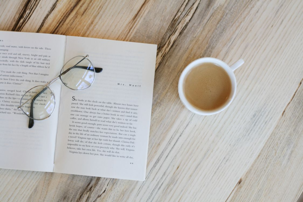 A minimalist flat lay arrangement of a coffee cup, an open book, and a pair of eyeglasses on a wooden table surface.