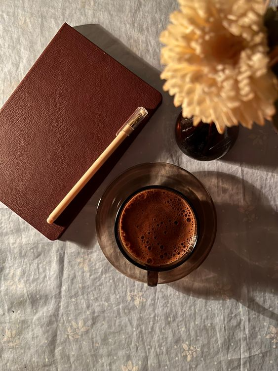 An elegant flat lay arrangement featuring a coffee cup, a journal with a pen, and a single brown flower on a textured wooden table.