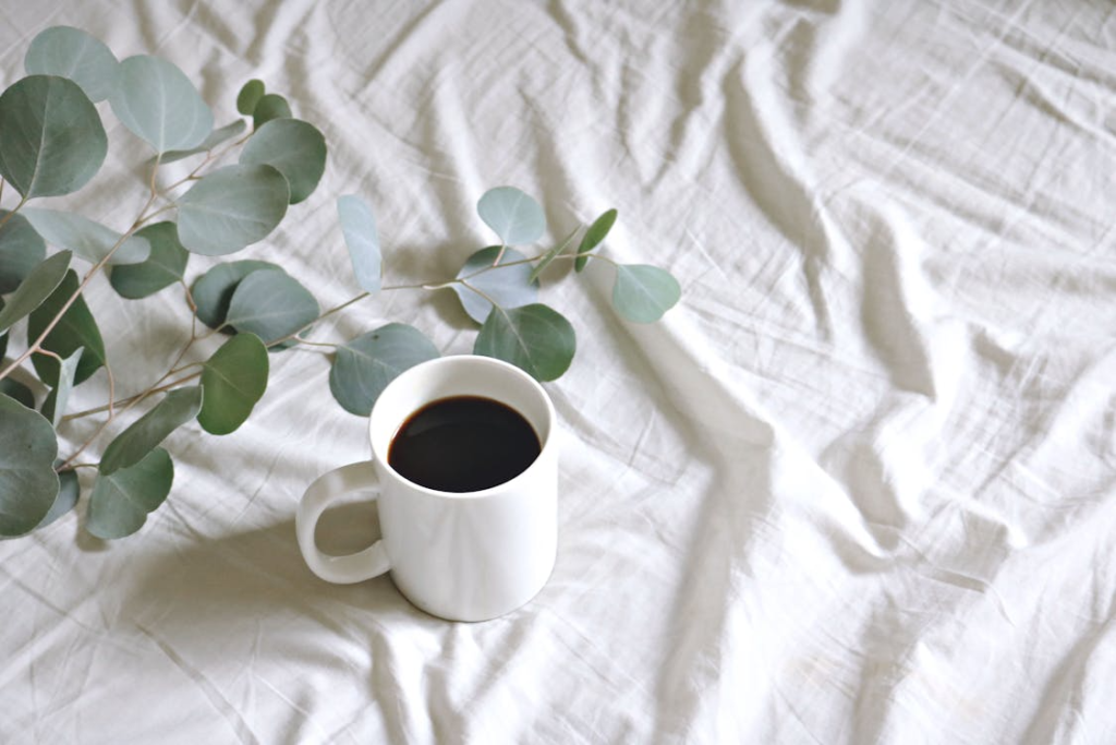 A simple white ceramic mug filled with coffee on a wooden surface beside a sprig of eucalyptus in soft natural light.