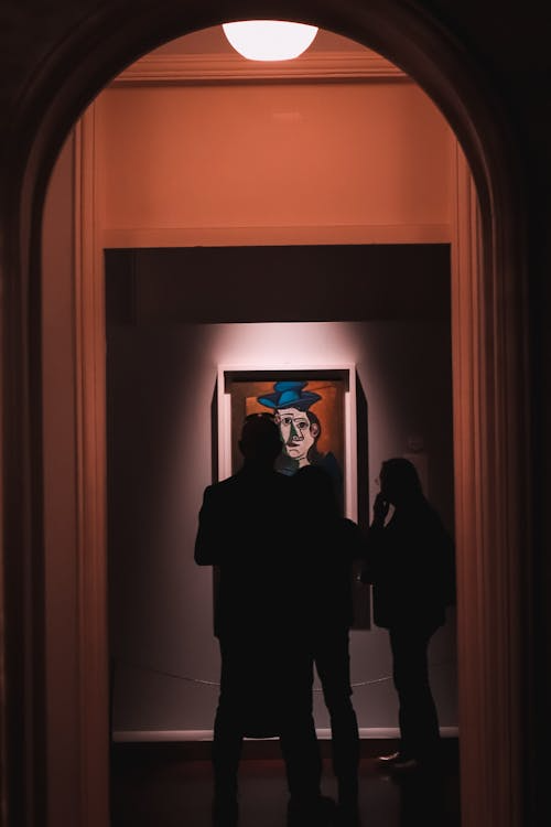 Silhouettes of people observing colorful abstract paintings in an art gallery corridor with dim lighting.