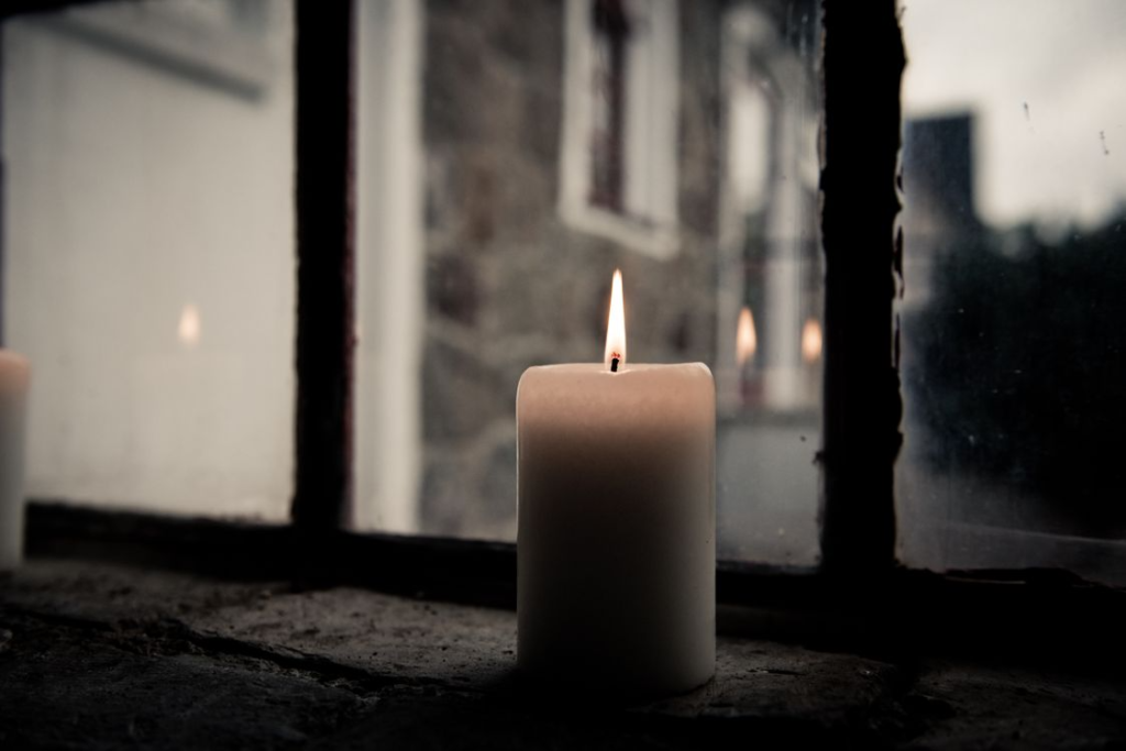 A lit pillar candle by a clear glass window, offering a warm, cozy, and contemplative ambiance.